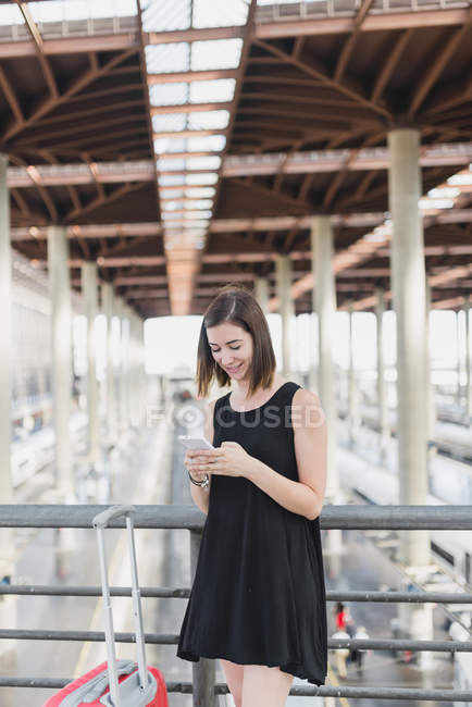 Portrait of brunette girl with red suitcase using smartphone at railway station — Stock Photo