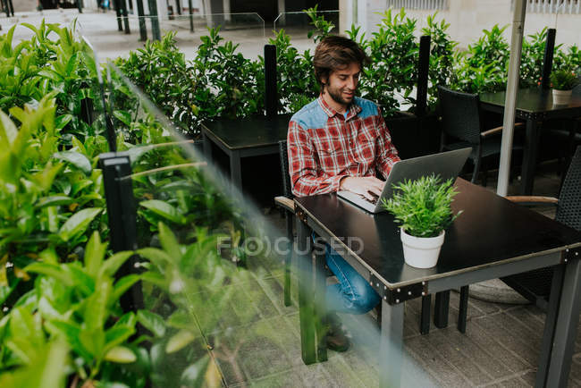 Jeune homme en chemise à carreaux tapant sur ordinateur portable tout en étant assis dans un café extérieur — Photo de stock