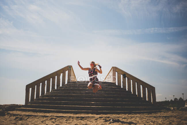 Молода дівчина в барвистій сукні і сонцезахисних окулярах стрибає на пляжі проти кам'яних сходів — стокове фото