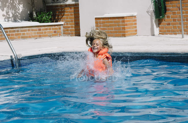 Niña feliz en chaleco salvavidas entrando al agua en la piscina con los ojos cerrados . - foto de stock