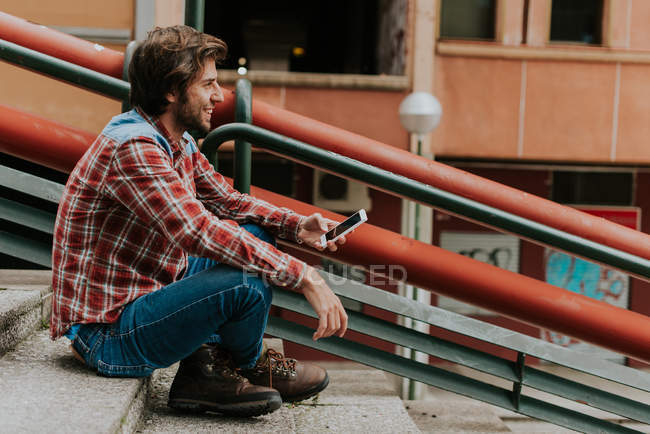 Sorrindo homem em passos com o smartphone na mão olhando para longe — Fotografia de Stock