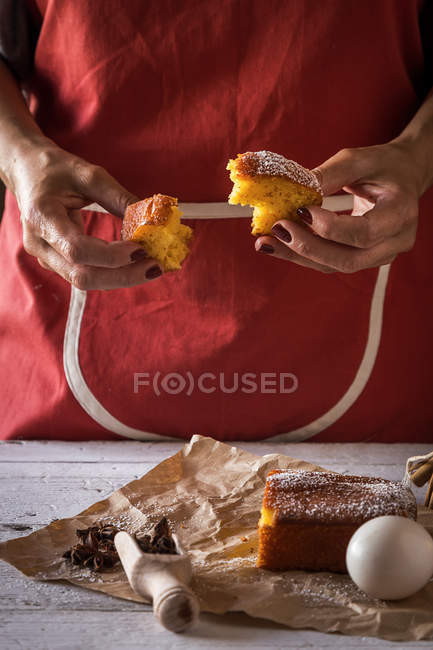 Partie médiane de la tranche de gâteau au citron déchirant femelle sur la table avec gâteau et ingrédients sur papier de boulangerie — Photo de stock