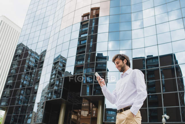 Vista lateral del hombre de negocios sonriente en camisa blanca usando el teléfono sobre la fachada del edificio del negocio en el telón de fondo - foto de stock