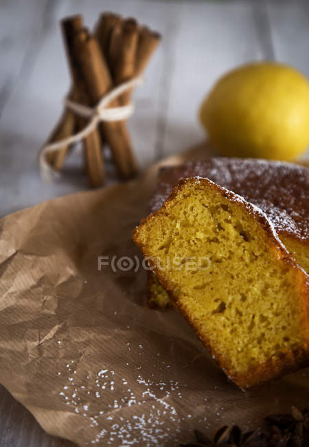 Close up view of homemade cake slices on bakery paper over cinnamon sticks and lemon on white wooden table on backdrop — Stock Photo