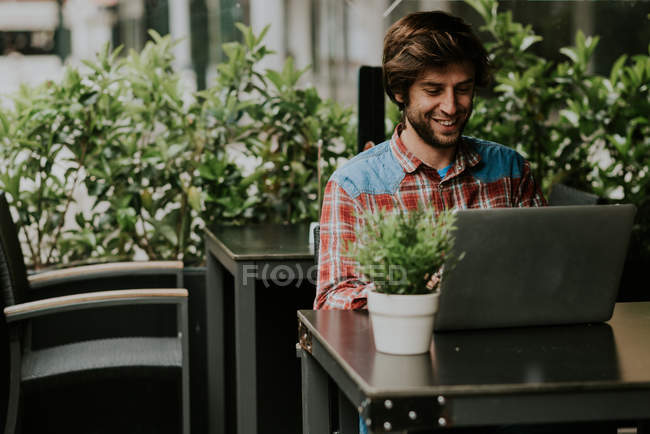 Portrait d'un homme barbu assis à la table de terrasse d'un café avec une plante en pot et utilisant un ordinateur portable — Photo de stock