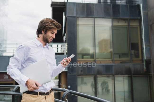 Side view of businessman carrying laptop and using smartphone over business building facade on backdrop — Stock Photo
