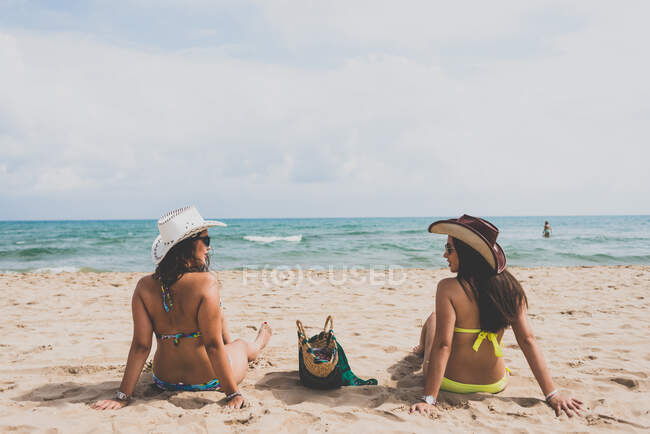 Back view of two young girlfriends in bikini and cowboy hats sitting on beach against wavy sea and cloudy sky. — Stock Photo
