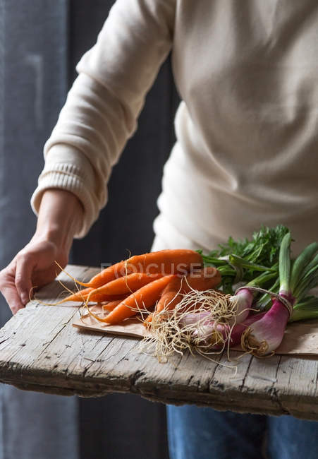 Mulher segurando cachos frescos de cenouras e cebolinha na tábua de madeira — Fotografia de Stock