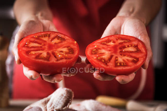 Primer plano de las manos femeninas sosteniendo tomate fresco a la mitad para preparar pollo - foto de stock