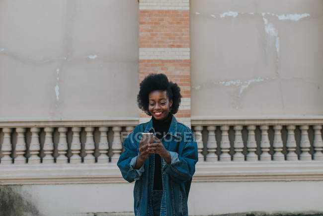 Улыбающаяся девушка в джинсовой одежде с помощью смартфона над старой грубой стеной — стоковое фото