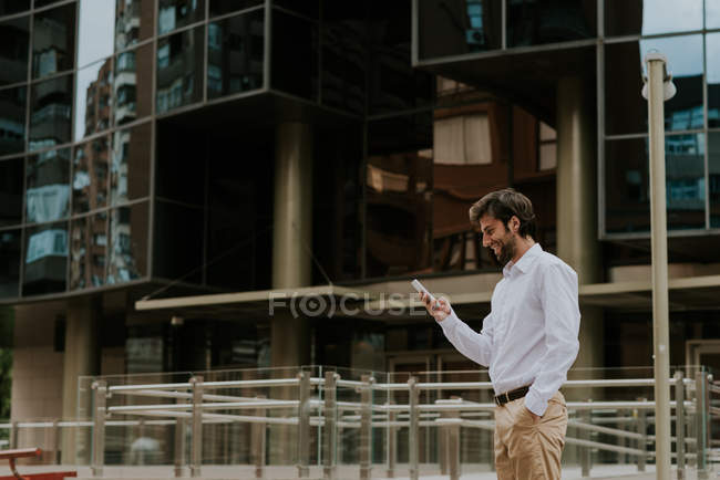Вид збоку усміхненого бізнесмена в білій сорочці, використовуючи телефон над фасадом бізнес-будівель на міській сцені — стокове фото