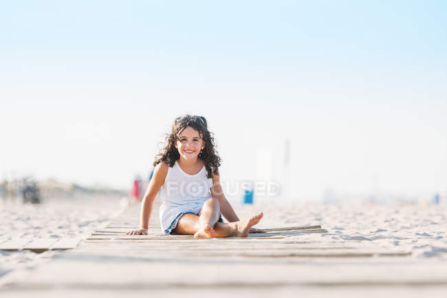 Retrato de chica alegre sentada en la arena en la playa y mirando a la cámara - foto de stock