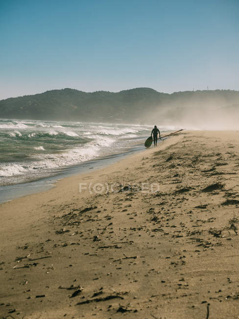 Persona con tabla que camina en la orilla arenosa con mareas fuertes que lavan la costa . - foto de stock
