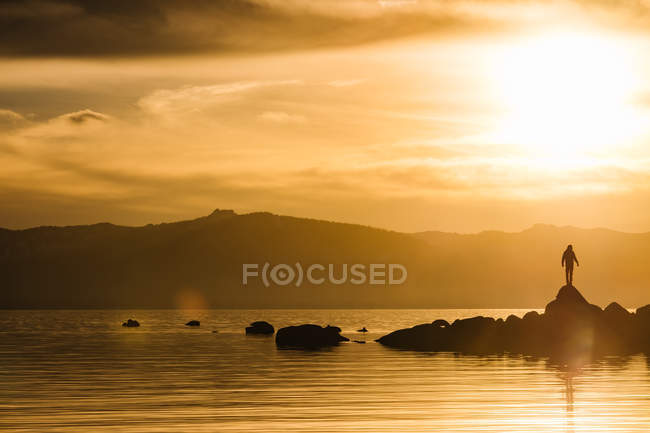 Silhouette of traveler on rock in water of lake on background of sunset sky. — Stock Photo