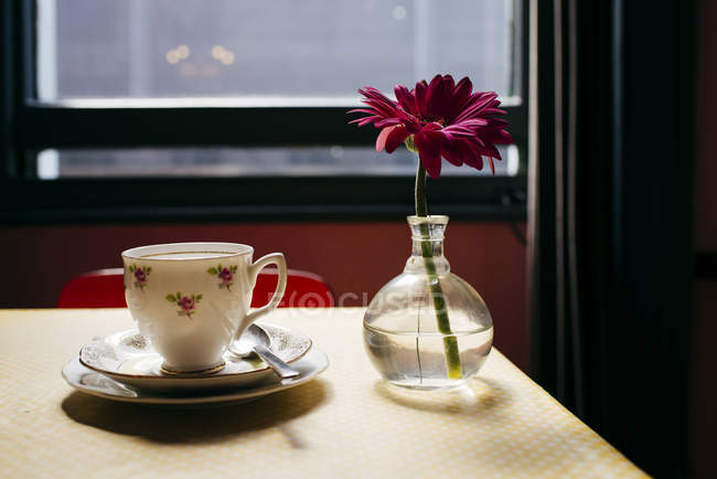 Still life of small glass vase with gerbera flower and cup of coffee on table. — Stock Photo