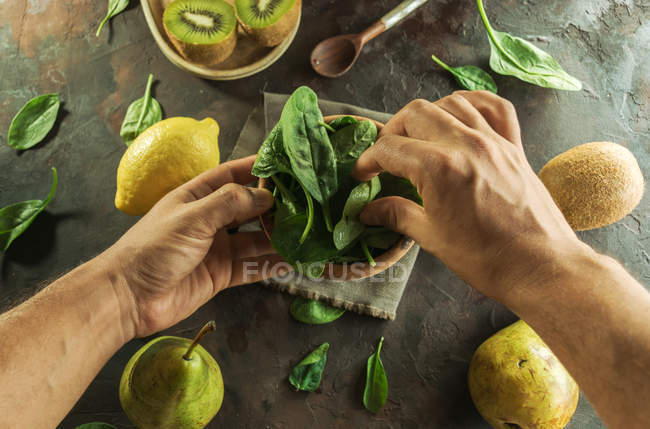 Vista superior de las manos recortadas sosteniendo un tazón de madera lleno de hojas de espinacas frescas en la mesa con - foto de stock