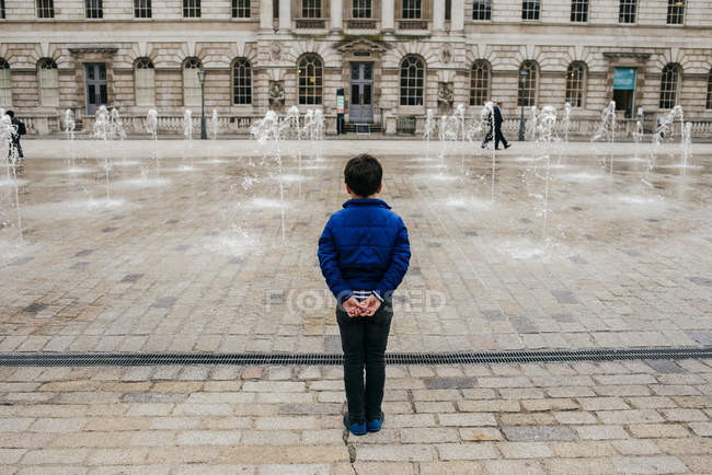 ЛОНДОН, Великобритания - 4 мая 2017 года: вид сзади маленького мальчика, стоящего перед площадью с фонтанами, брызгающими водой . — стоковое фото