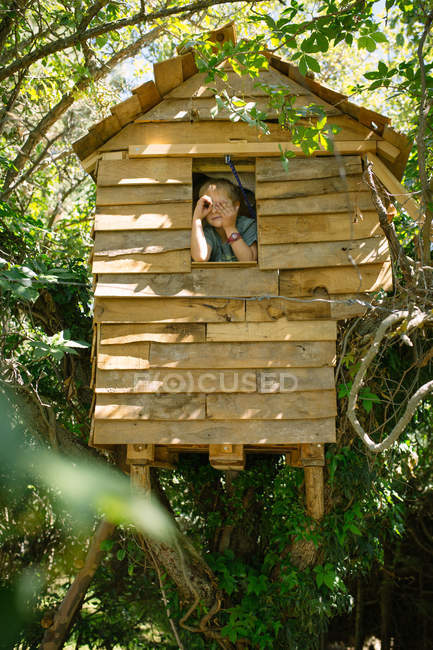 Bottom view of blonde boy looking out of wooden treehouse window and posing like using spyglass — Stock Photo