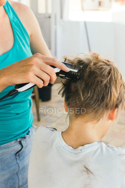 Imagen recortada de la madre cortando el pelo del hijo con la máquina eléctrica - foto de stock