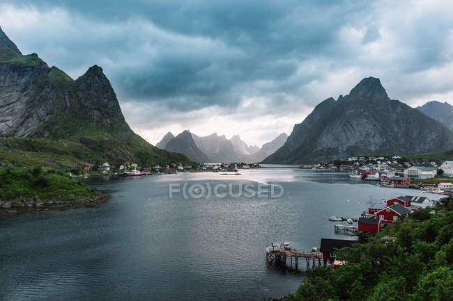 View of rocky mountains and lagoon with village on shores under gloomy sky. — Stock Photo