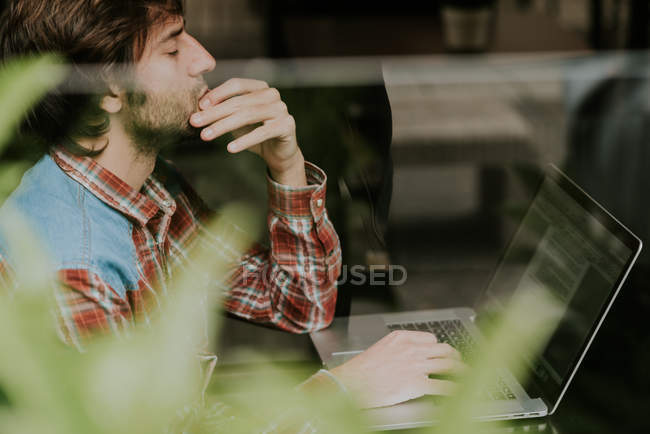 Вид збоку бородатого чоловіка в картатій сорочці з ноутбуком на терасі відкритого кафе — стокове фото