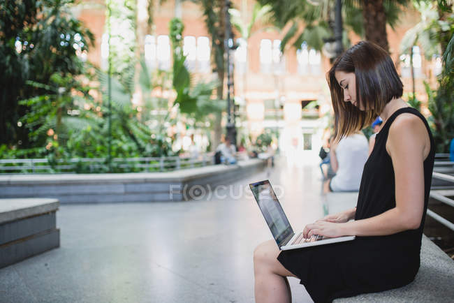 Vista lateral da jovem morena no vestido preto sentado e usando laptop nos joelhos — Fotografia de Stock