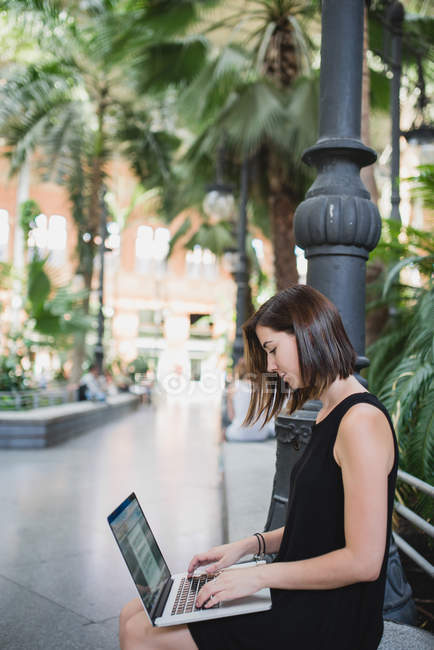 Vista laterale della giovane ragazza bruna in abito nero seduta e usando il computer portatile sulle ginocchia — Foto stock