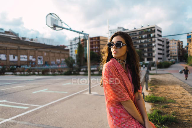 Brünettes Mädchen mit Sonnenbrille sitzt auf Zaun am Straßenspielplatz und schaut über die Schulter weg — Stockfoto