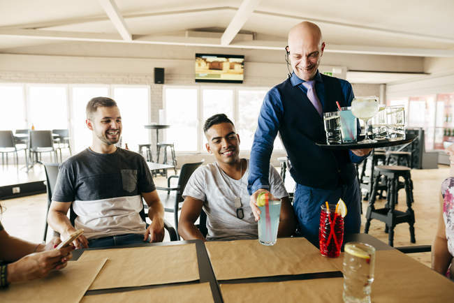 Homem garçom alegre servindo bebidas para amigos à mesa no bar . — Fotografia de Stock