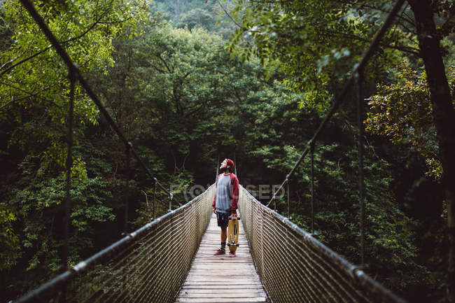 Человек со скейтбордом позирует на веревочном мосту в лесу — стоковое фото