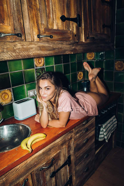 Чувственная женщина лежит на кухонном столе с посудой и бананами и смотрит в камеру — стоковое фото