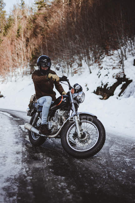 Mann mit Helm posiert auf Chrom-Motorrad mit Hintergrund kahler Bäume auf schneebedeckter Straße. — Stockfoto