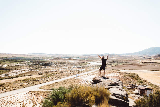 Rear view of cheerful man standing on cliff with raising arms with camera — Stock Photo