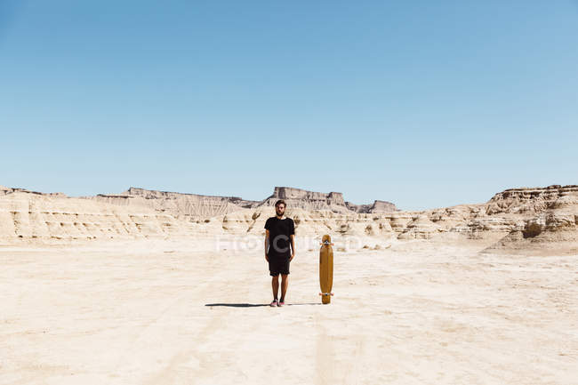 Uomo in abiti neri in piedi vicino skateboard al deserto — Foto stock