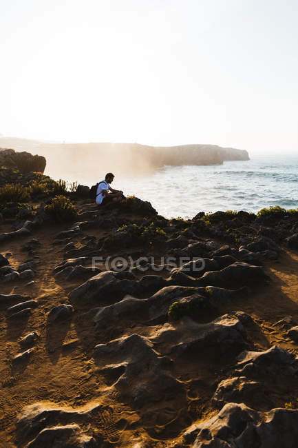 Hombre sentado en el acantilado sobre el paisaje costero - foto de stock