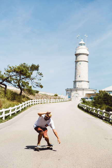 Side view of skateboarder man taking a ride on asphalt alley to lighthouse. — Stock Photo
