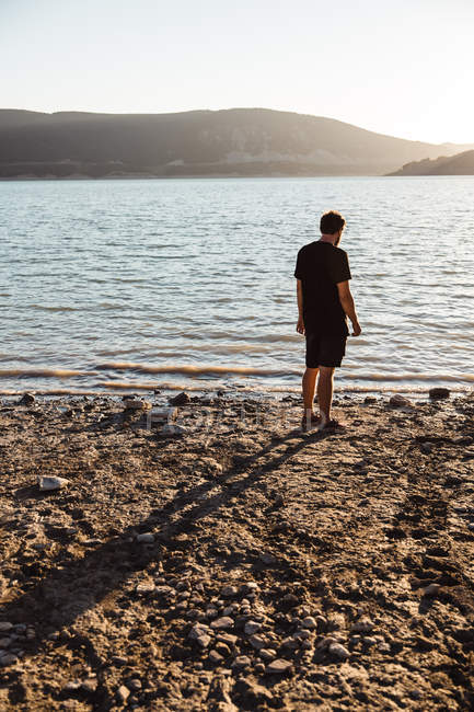 Vue arrière de l'homme en posant sur la plage sur une colline brumeuse à la rive opposée en arrière-plan — Photo de stock