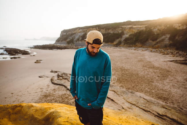 Hombre barbudo en suéter y gorra de pie mirando hacia abajo en el acantilado en la playa de arena - foto de stock