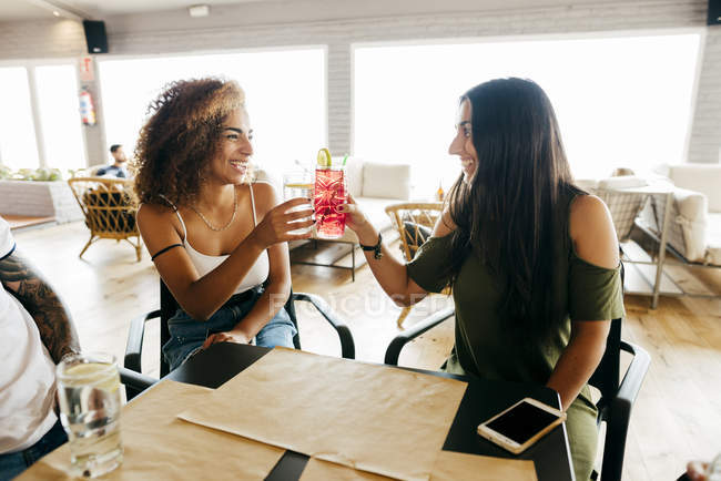 Two women sitting at table in cafe and clinking cocktails — Stock Photo