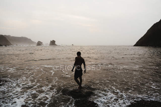 Вид сзади на человека, идущего в волнах серфинга на туманном берегу моря — стоковое фото
