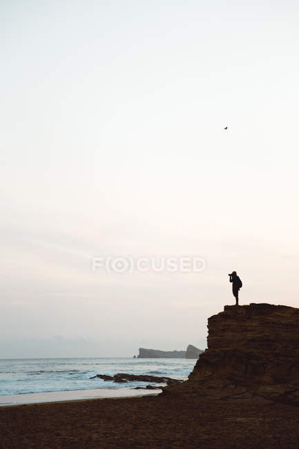 Vista lateral de la silueta del hombre de pie en el acantilado costero y tomar fotos con cámara - foto de stock