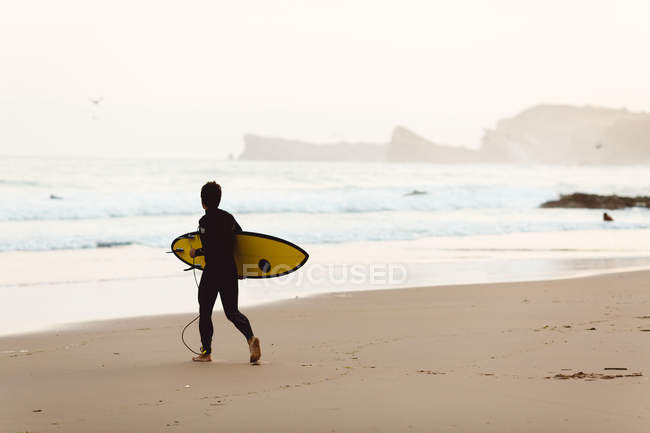 Rear view of child surfer walking to waves on sandy shore at seaside. — Stock Photo