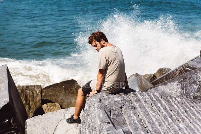 Задний вид человека, сидящего у камней возле брызг волн на море и оглядывающегося через плечо — стоковое фото