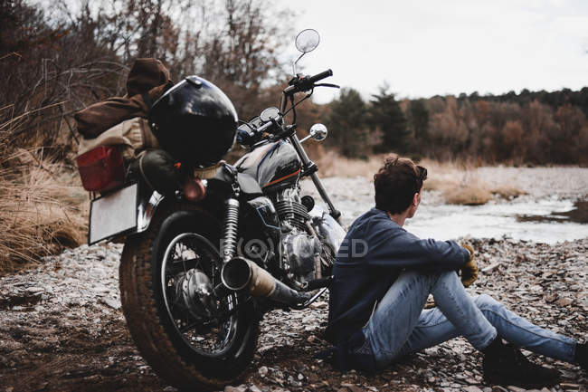 Hombre sentado junto a la motocicleta estacionada y admirando la orilla del río guijarro - foto de stock