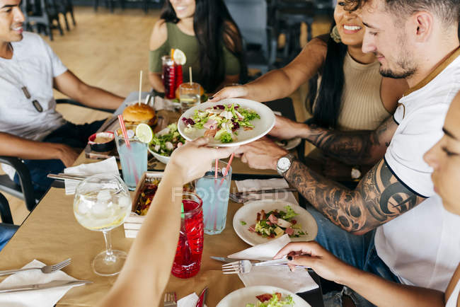 Amigos pasando plato de ensalada mientras están sentados y comiendo juntos en el restaurante . - foto de stock