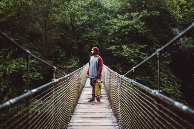 Vue latérale de l'homme avec planche à roulettes posant sur un pont de corde forestière — Photo de stock