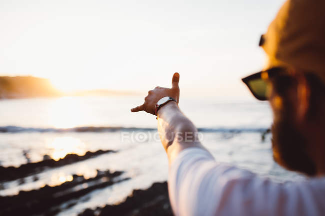 Hombre de las cosechas en gafas de sol haciendo gestos sobre la puesta de sol junto al mar en el fondo - foto de stock