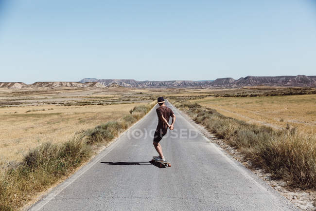 Vista posteriore di uomo locanda cap equitazione skateboard su strada prateria — Foto stock