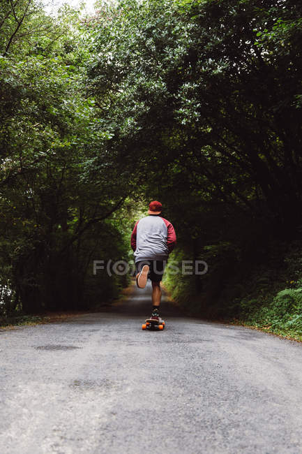 Visão traseira do homem no skate montando na estrada da floresta — Fotografia de Stock