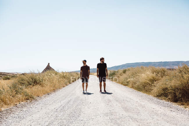 Dois homens de t-shirts e calções andando ao longo da estrada da pradaria no dia ensolarado. — Fotografia de Stock